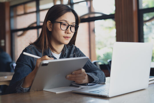 Focused woman using digital tablet and stylus with laptop computer in modern office, studying or working with digital devices.