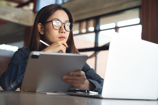 Woman using digital tablet and stylus thinking and looking outside with laptop computer on desk in cafe modern workspace, working or studying with digital technology.