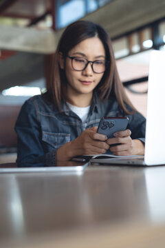 Young asian woman using smartphone during working on laptop at desk in modern workspace, remote working in coffee shop, modern business lifestyle. vertical