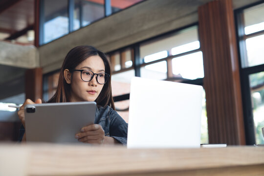 Focused woman using digital tablet and stylus, looking at  laptop screen, remote working in modern office, studying or working with digital devices.