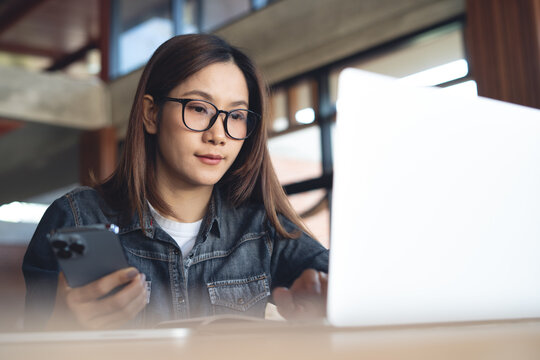 Young asian woman online working on laptop, hand holding smartphone at desk in modern workspace, remote working in coffee shop, online business lifestyle.