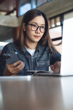 Young asian woman online working on laptop, hand holding smartphone at desk in modern workspace, remote working in coffee shop, online business lifestyle. vertical