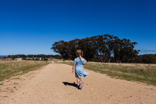 Little farm girl under blue summer sky running down gravel road through paddocks