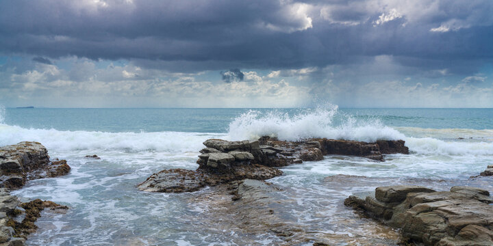 Panorama view of waves crashing over shoreline rocks under dark clouds