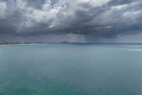 Aerial view of rain falling from dark clouds over a calm turquoise sea and distance hills