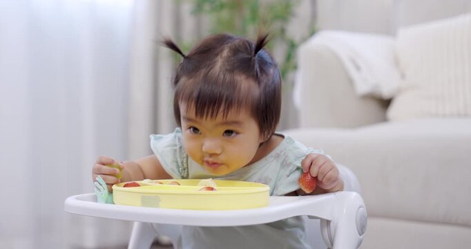 Asian toddler girl during mealtime holds strawberry piece, chews gently, looks thoughtful, small hand movement natural, cozy living room background, calm color tone, early self feeding, child