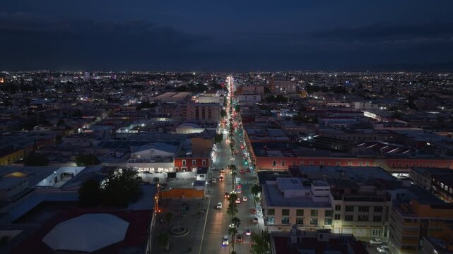 Aerial drone footage of Durango Mexico cityscape at night showing busy main street with car traffic and glowing streetlights. Urban landscape extends to horizon under dramatic cloudy evening sky.