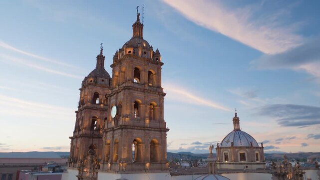 Aerial drone footage slowly pans across magnificent Durango Cathedral featuring ornate baroque twin bell towers with prominent clock face and beautiful blue-tiled dome against stunning sunset sky.