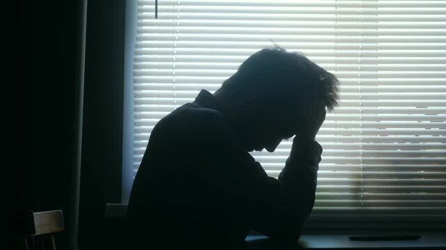 Sad and depressed man in silhouette sits alone at table in dark room, his head in his hands, representing despair, anxiety, loneliness, mental health issues. Depressed, sad man sitting in dark room