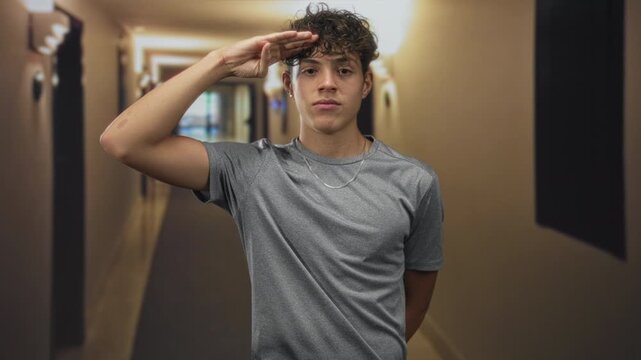 Teenage man salutes with right hand revealing forearm and chain necklace in dim hotel building corridor; respectful.