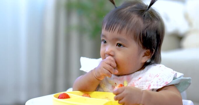 Asian toddler girl eating fruit with fingers on feeding tray, smiling softly with happiness, learning to self feed through play, showing curiosity and joy, warm indoor family moment, early childhood