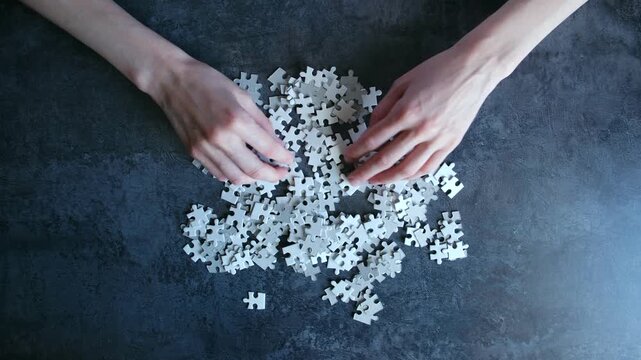 Female hands mixing, sorting pile of blank white jigsaw puzzle pieces on dark, textured tabletop, representing complex problem, challenge, beginning of new project. Person mixing white jigsaw puzzle