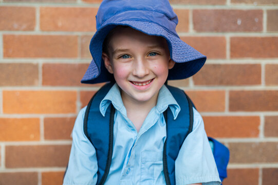 Boy in a school uniform with a blue hat sits against a brick wall and smiles