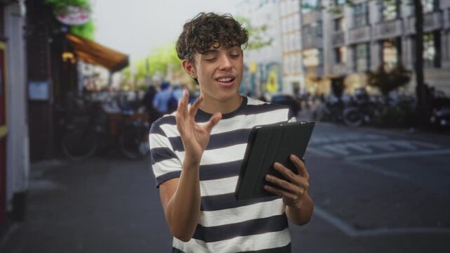 Teen man in striped shirt holding tablet and making ok gesture on a busy street cafe, smiling and reading from screen; joy curiosity.