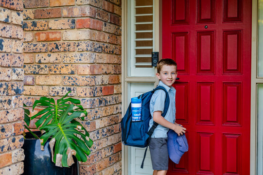 Boy with a school backpack stands at the red front door of his home