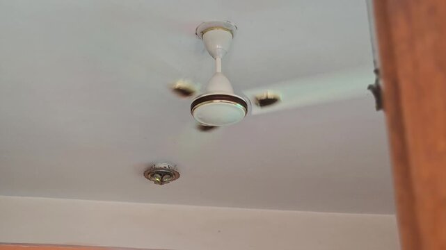 A white ceiling fan rotates slowly, its gentle motion struggling against the sweltering indoor heat. Blurred blades and shimmering air capture the oppressive atmosphere of a summer temperature peak.