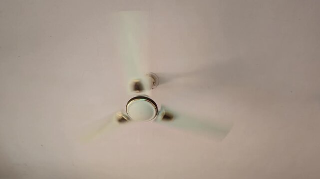 A white ceiling fan spins slowly against a pale ceiling, evoking a stifling summer day. Shimmering heat waves suggest high temperatures and the struggle for relief through a gentle, rhythmic breeze.