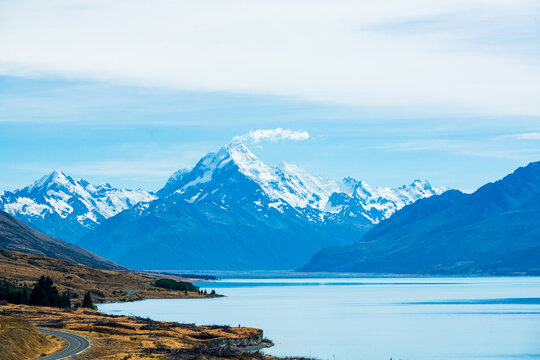 View of road alongside a tranquil blue lake to alpine snow capped mountains with clouds and blue sky