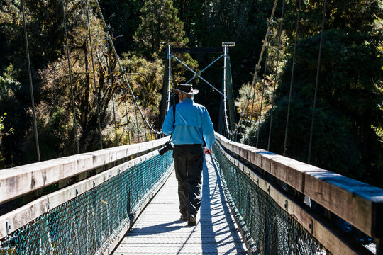 Back view of older man wearing hat and with camera walking along swing bridge with sharp shadows