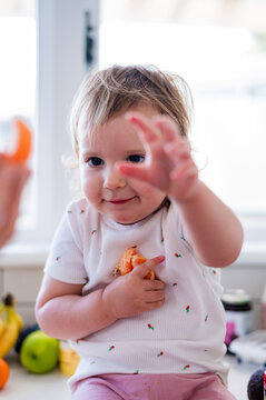 Toddler girl sitting on kitchen counter holding peeled mandarin wedges with other hand reaching out