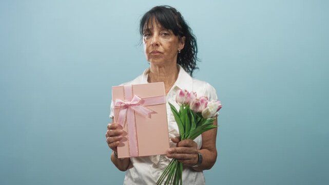 Senior hispanic woman holding a pink gift box tied with ribbon and a bouquet of pink and white tulips, hands visible and wearing white blouse in studio; melancholy reflection.