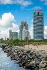 Fototapeta premium Vertical view of rock jetty and Miami skyscrapers. Large stones of a jetty leading towards luxury residential towers under a blue sky in Miami Beach.