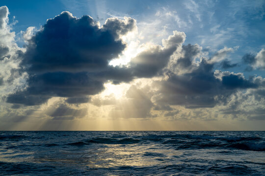 Sunlight through heavy clouds over Atlantic Ocean at dawn. Intense sun rays breaking through grey clouds over sea water at Miami Beach, USA.