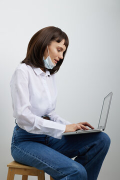 A focused woman sits on a wooden stool, wearing a light blouse and jeans, typing on a laptop with a mask tucked under her chin, presenting a calm home workspace.