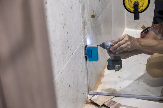 Focused construction worker install transparent glass shower room divider using electric power drill tool on tile wall during modern bathroom renovation and home improvement project