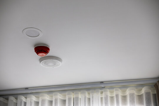 Red fire alarm smoke detector and white recessed led downlight fixture installed on residential ceiling providing reassuring safety and modern illumination in comfortable home interior