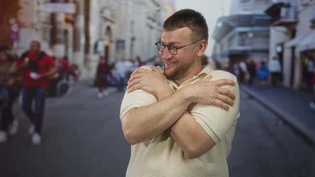 Man smiling and reaching forward then hugging his forearms with crossed arms on a busy city street in daytime; self care joy.