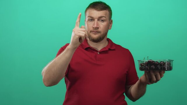 Man points finger to head while holding a clear punnet of cherries in a teal green studio; thinking decision.