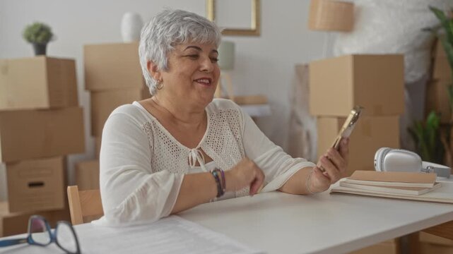 Woman holding smartphone, covering mouth and waving inside a building with stacked moving boxes and notebooks on table; joy connection.