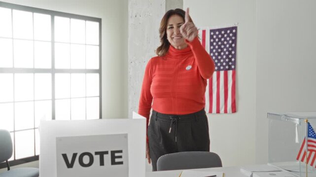 Woman giving thumbs up and pointing finger at ballot box and vote booth in a polling building; civic pride.