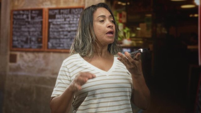 Hispanic woman holding smartphone and gesturing while speaking into phone in a building cafe interior with chalk menu and counter; frustration.