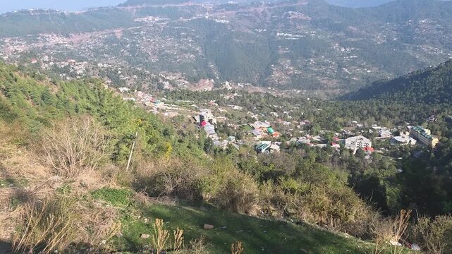 High-Angle View of a Hill Station Village Nestled in Green Mountains. A wide high-angle shot overlooking a scenic valley in a mountain range. The frame shows traditional houses and buildings scattered