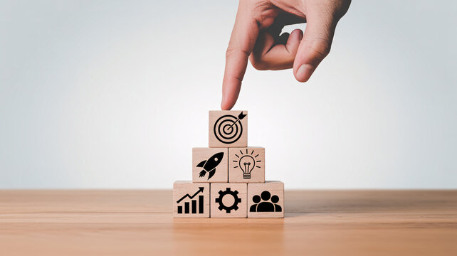 A hand placing a wooden block with a target icon on top of a stack of blocks with various business and strategy symbols.