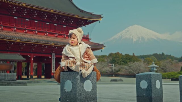 Adorable Asian toddler girl sitting on a stone pillar at a Japanese temple with Mount Fuji background. Happy child enjoying winter family vacation and cultural sightseeing in Japan, expressing joy