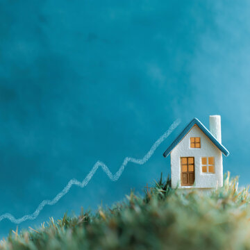 Rising Real Estate: A miniature house sits atop a grassy hill with a upward trending graph in the background, symbolizing the increase in housing value and the growth of the property market.