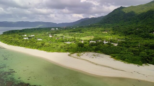 Dramatic drone panorama of Sukuji Beach's white sands and emerald reefs, transitioning to the towering, lush green mountain peaks of Ishigaki Island, Okinawa