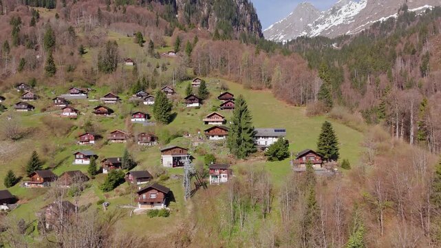 Alpine hamlet of Schlattberg above Luchsingen, Switzerland with traditional chalets on a hillside and snow-capped mountains in background. Aerial pull back.