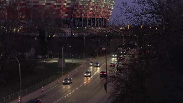 Evening traffic flowing on wybrzeze szczecinskie street in warsaw, with the iconic illuminated pge narodowy football stadium in the background