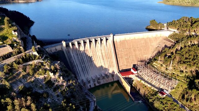 Smooth drone pull away shot from Presa de Izn&aacute;jar revealing concrete dam, blue reservoir, and surrounding hills in Andalusia, Spain, highlighting infrastructure and water management.