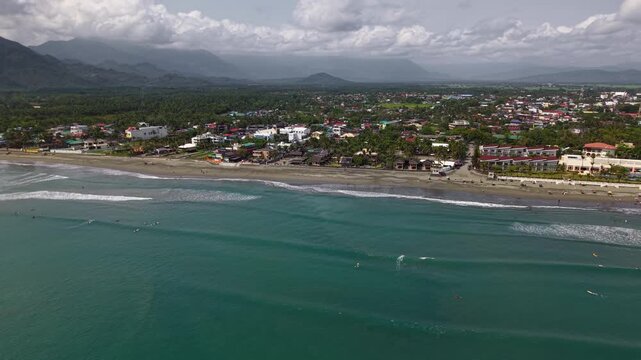 Scenic aerial drone video of Baler Beach in the Philippines captures surfers riding waves with mountains in the background and coastline under a bright tropical sun.