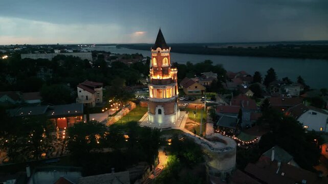 Dramatic drone orbit around illuminated Gardos tower at twilight with incoming lightning storm over Danube river. Storm clouds and lightning striking the background over Zemun, Belgrade, Serbia.