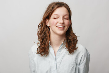 Obraz premium Young woman with curly hair smiles with closed eyes wearing white shirt on light background. Concept of natural beauty, happiness, casual style, and relaxed mood in studio portrait.