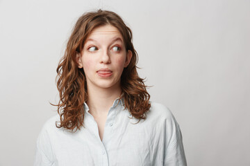 Obraz premium Young woman with curly hair wearing white shirt looks sideways with playful expression sticking out tongue. Studio portrait on light background. Concept of fun, youth, and casual style.