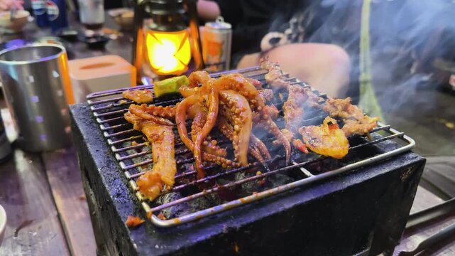 Close-up of marinated octopus tentacles and meat sizzling on a tabletop charcoal grill. Authentic Vietnamese BBQ experience during a river boat cruise in Hoi An, Vietnam. Vibrant food scene