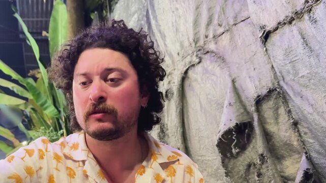 Portrait of a young man with curly dark hair and a mustache wearing a tropical palm tree print shirt. Candid indoor shot with a neutral textured background and green plants