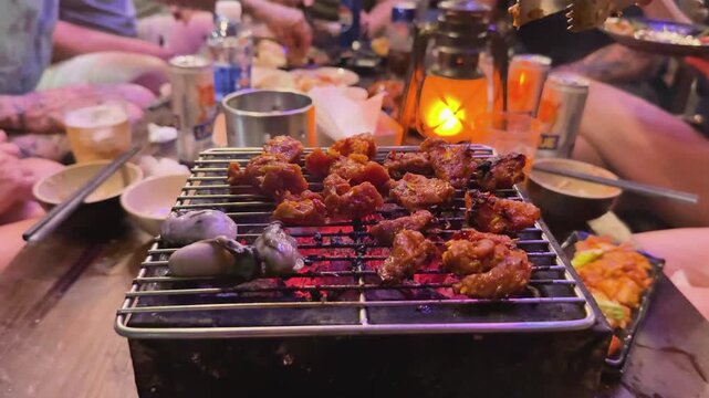 Slow motion close-up of traditional Vietnamese BBQ meat and seafood grilling on a charcoal brazier. Tongs flipping marinated pork at a vibrant night market street food stall in Vietnam